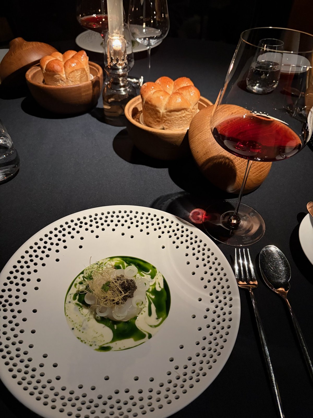 Dinner at Aska: a plated dish with green sauce, bread, and red wine on a dark table.